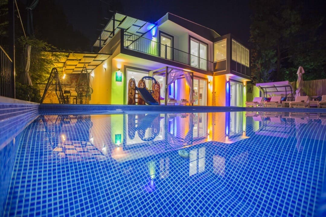 Gece ışıklandırılmış, havuzlu ve bahçesinde çocuk kaydırağı bulunan iki katlı modern bir villa.