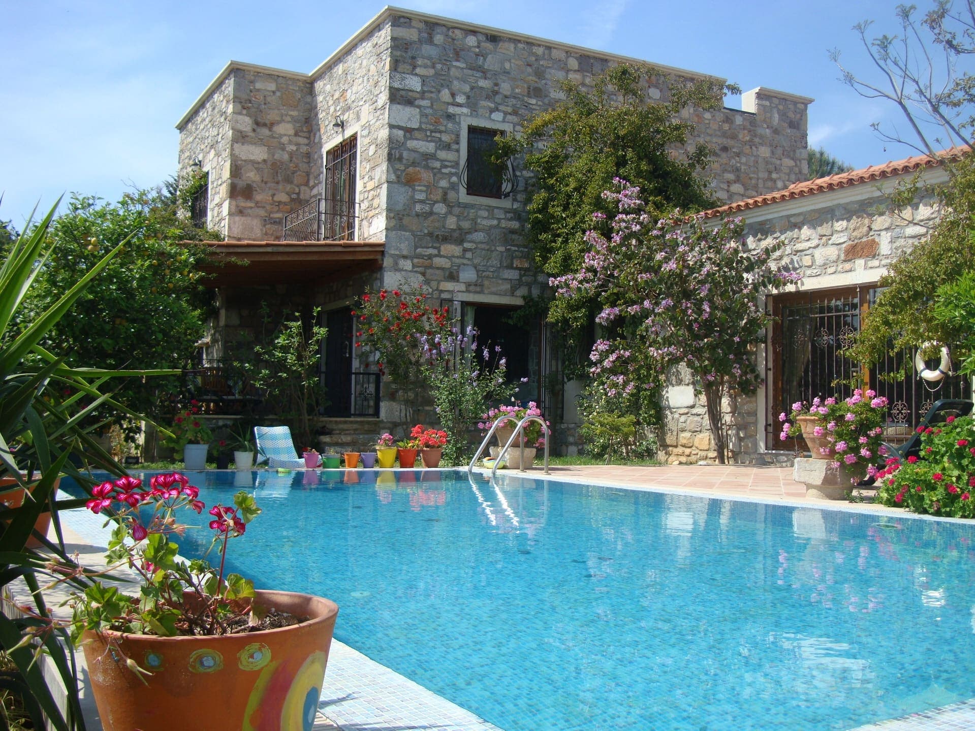Taş cepheli iki katlı villa önünde çiçekli havuz ve güneşlenme terası bulunuyor.
