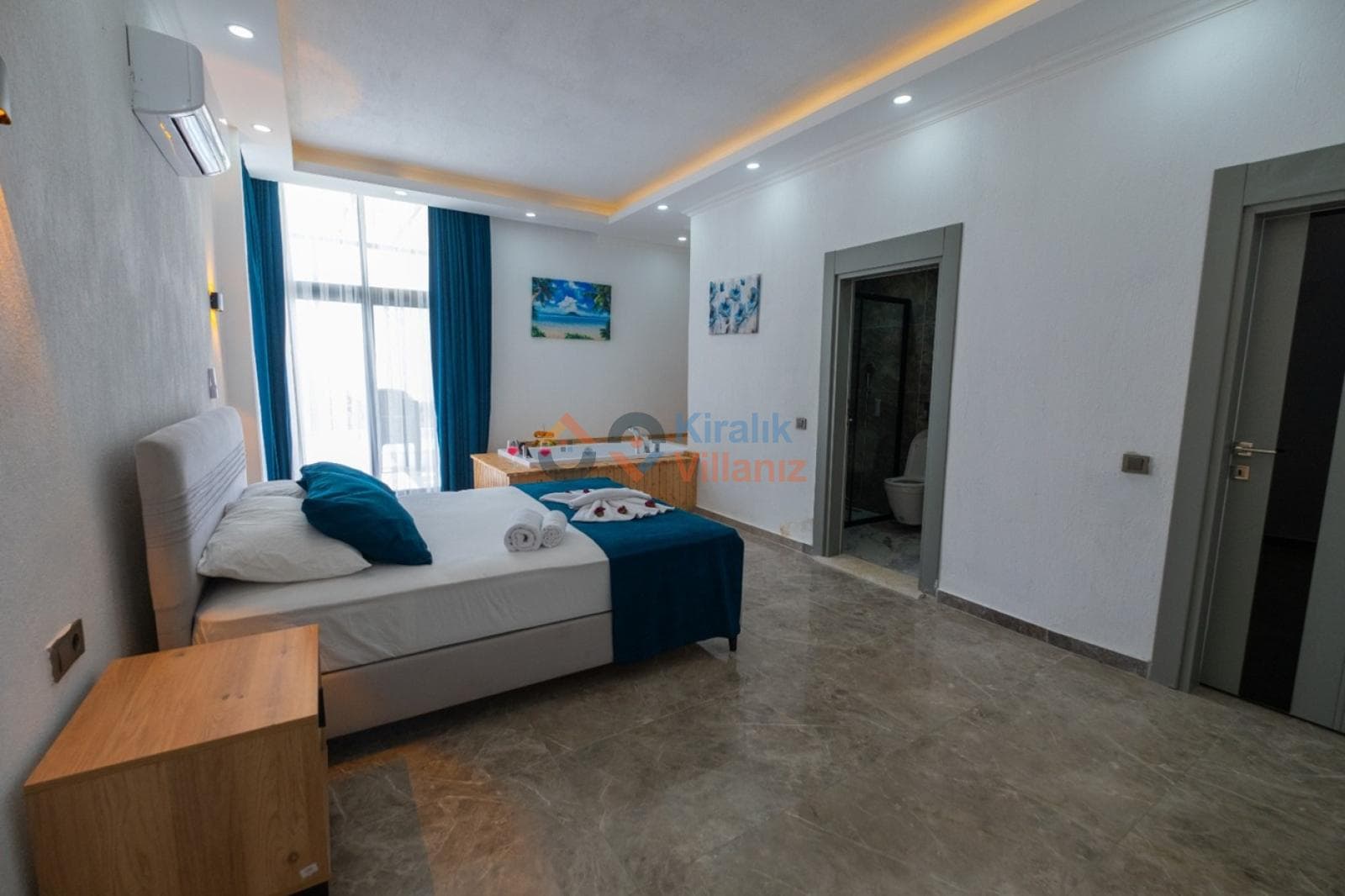 Mavi tonlarda yatak odasında ahşap komodin, jakuzi ve açık kapılı banyo yer alıyor.