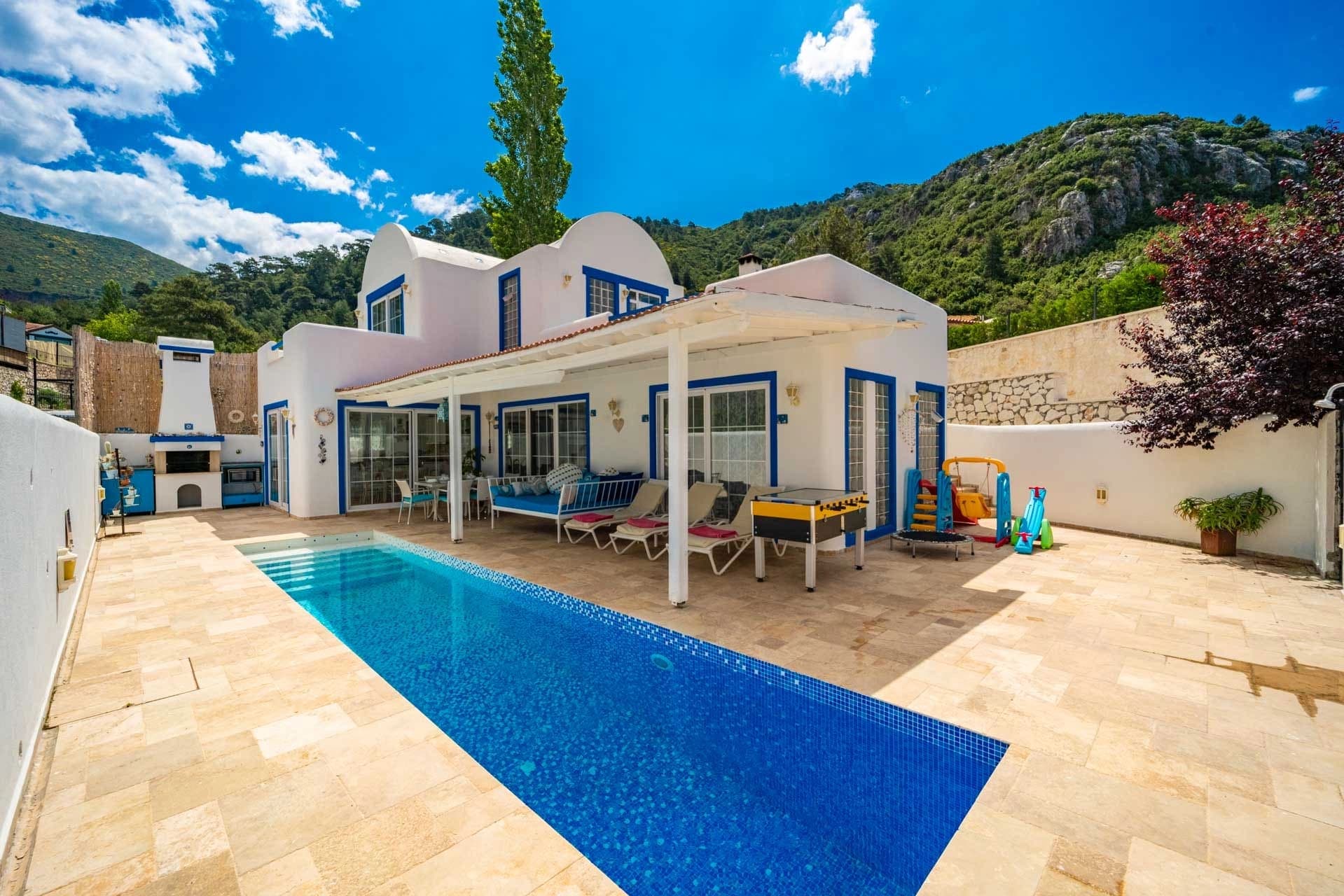 Mavi çerçeveli pencereleri olan beyaz villa ve önündeki uzun havuz.