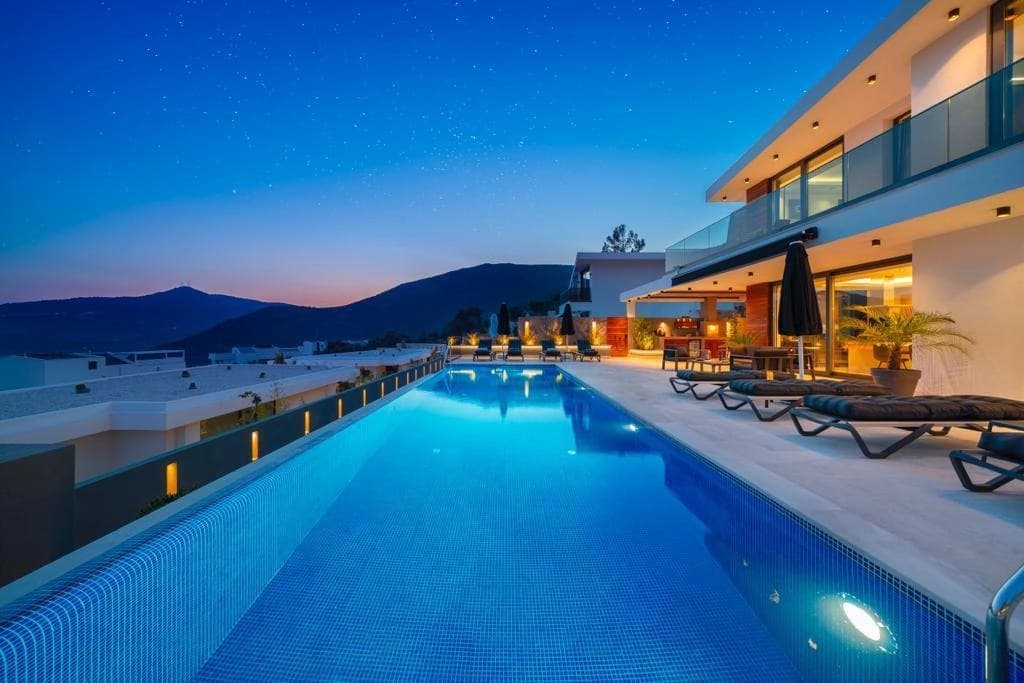 Akşam karanlığında dağ manzaralı havuz kenarı ve aydınlatmalı villa dış cephesi.