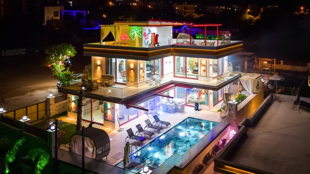 Gece ışıklandırılmış, havuzlu, teraslı ve Villa Red Palm neon tabelası bulunan üç katlı modern villa.