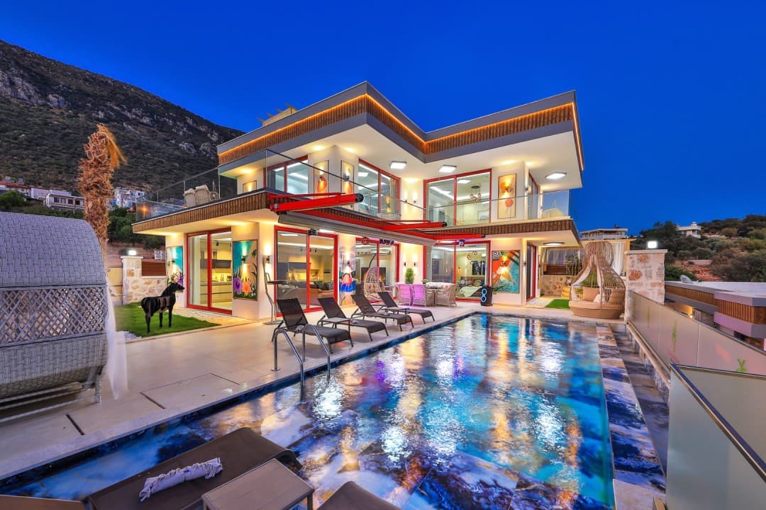 Gece ışıklandırmalı modern villa, önünde havuz ve şezlonglar.