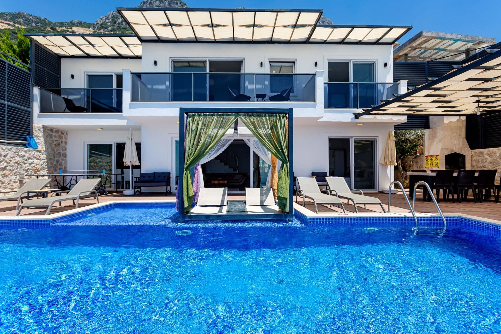 Güneşlikli havuz başı şezlongları ve beyaz dış cepheli villa.