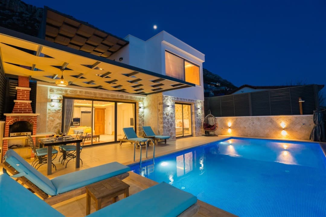 Akşam vakti havuz kenarında şezlonglar, yemek masası ve taş detaylı iki katlı villa.