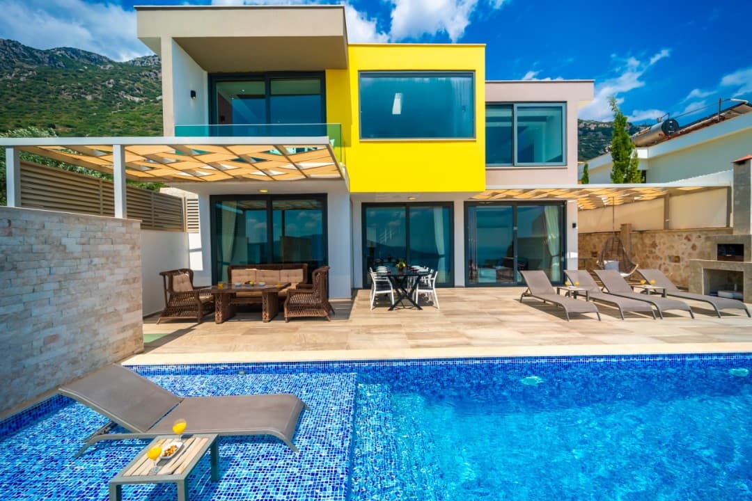Sarı ve beyaz dış cepheli, havuz başında şezlonglar, oturma grubu ve yemek masası bulunan modern villa.