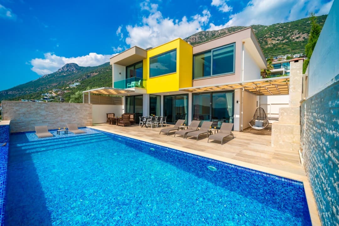 Önünde havuz ve şezlonglar bulunan, dağ manzaralı modern villa dış cephesi.