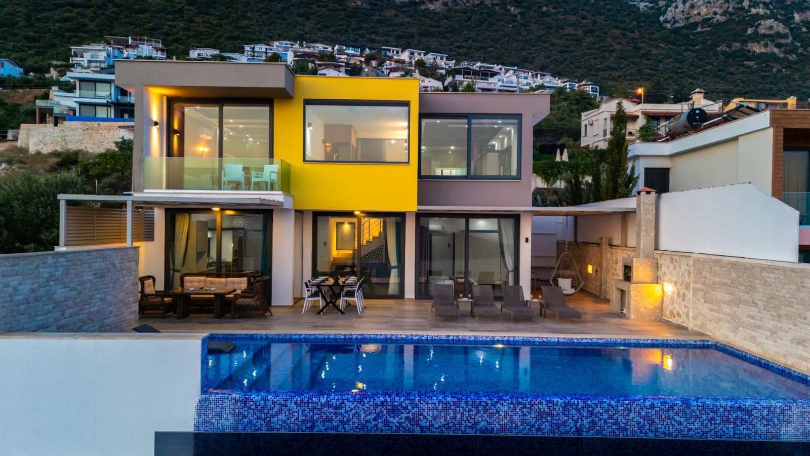 Akşam ışıklandırılmış havuz ve sarı cepheli modern villanın dış görünümü.