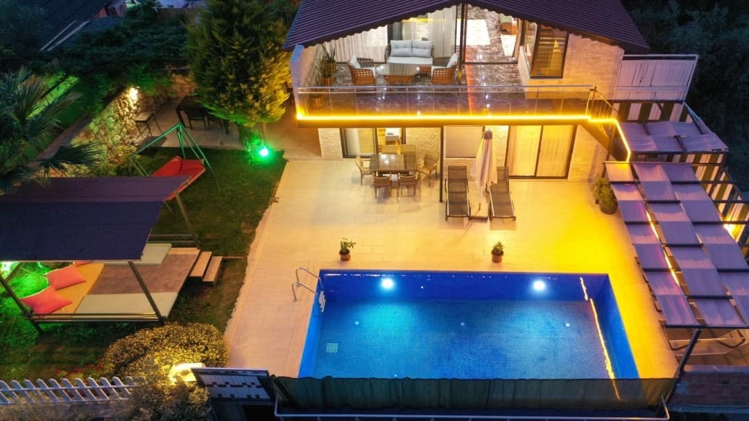 Aydınlatmalı havuzu, şezlongları ve çardaklı bahçesi olan iki katlı villa.