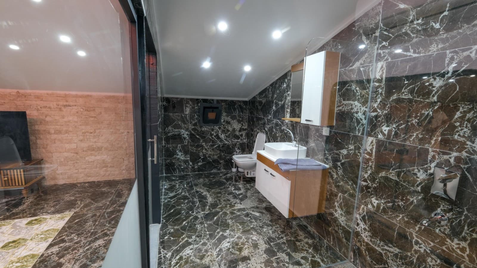 Siyah mermer duvar ve zemin kaplamalı, cam duş kabini ve beyaz lavabolu banyo.
