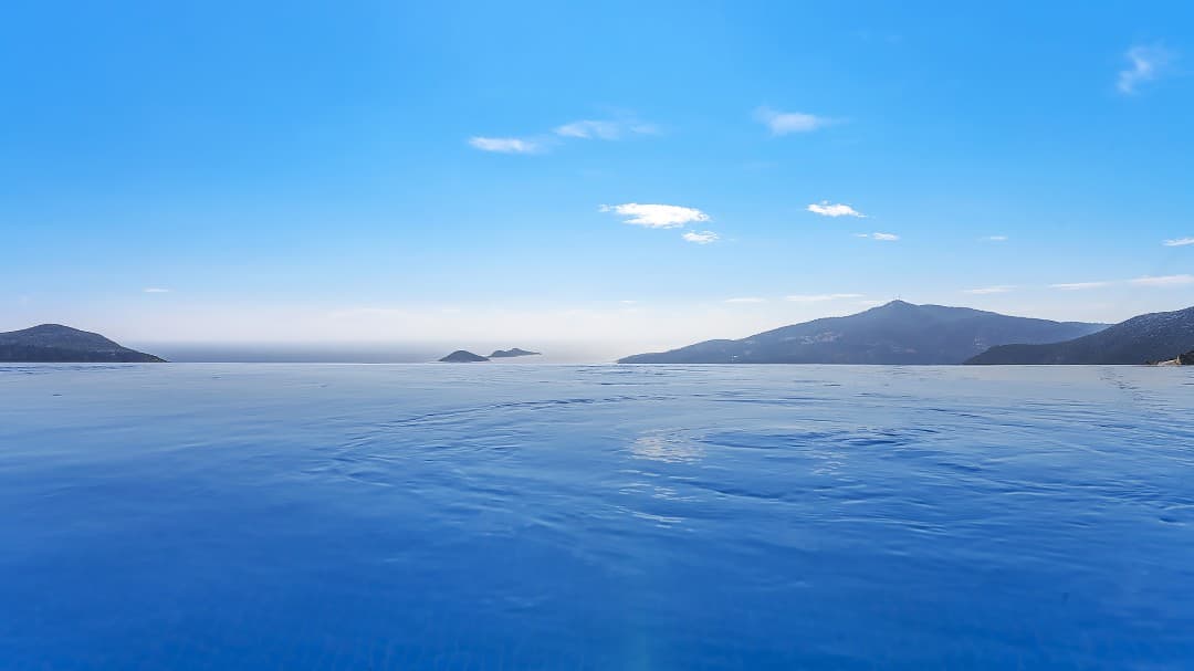 Açık mavi havuzun üzerinden ufuktaki deniz ve dağ manzarası.