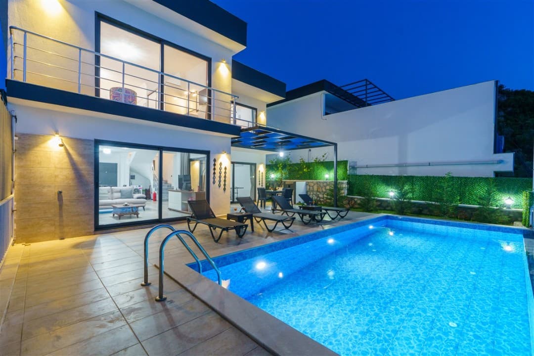 Akşam vakti aydınlatılmış, havuz başında şezlongların bulunduğu iki katlı modern villa.