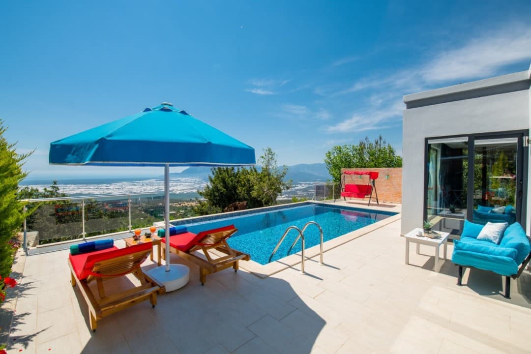 Mavi şemsiyeli havuz kenarında kırmızı şezlonglar, bir salıncak ve vadi manzaralı modern bir villa terası.