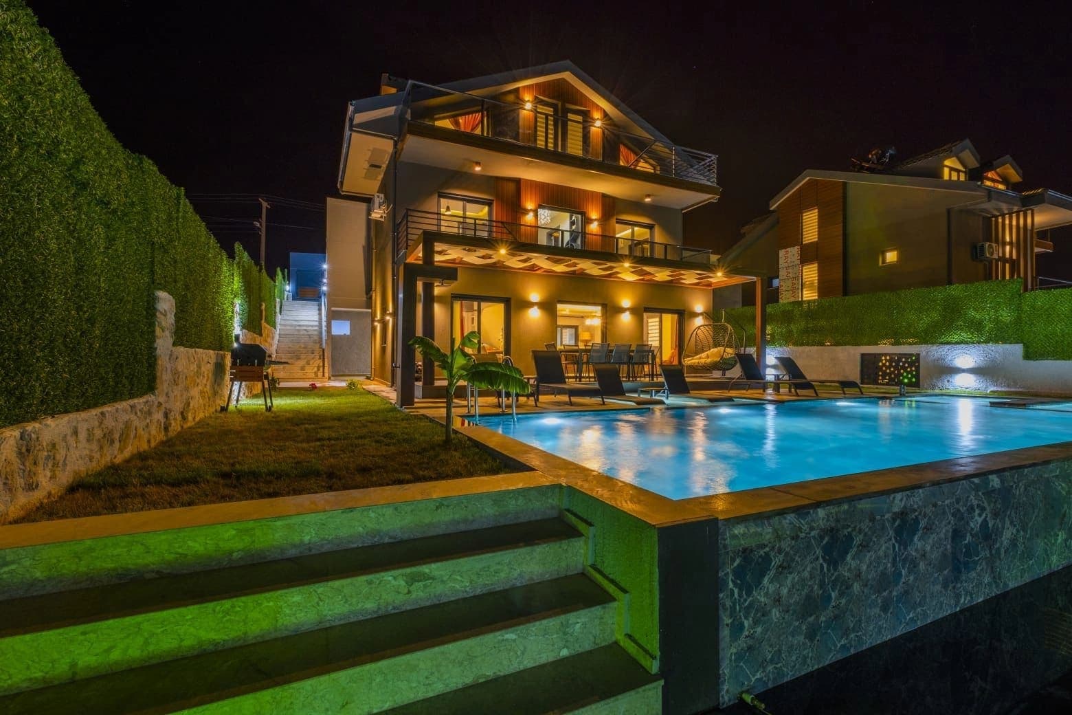 Gece ışıklandırmalı havuz kenarında şezlonglar ve arka planda villa.