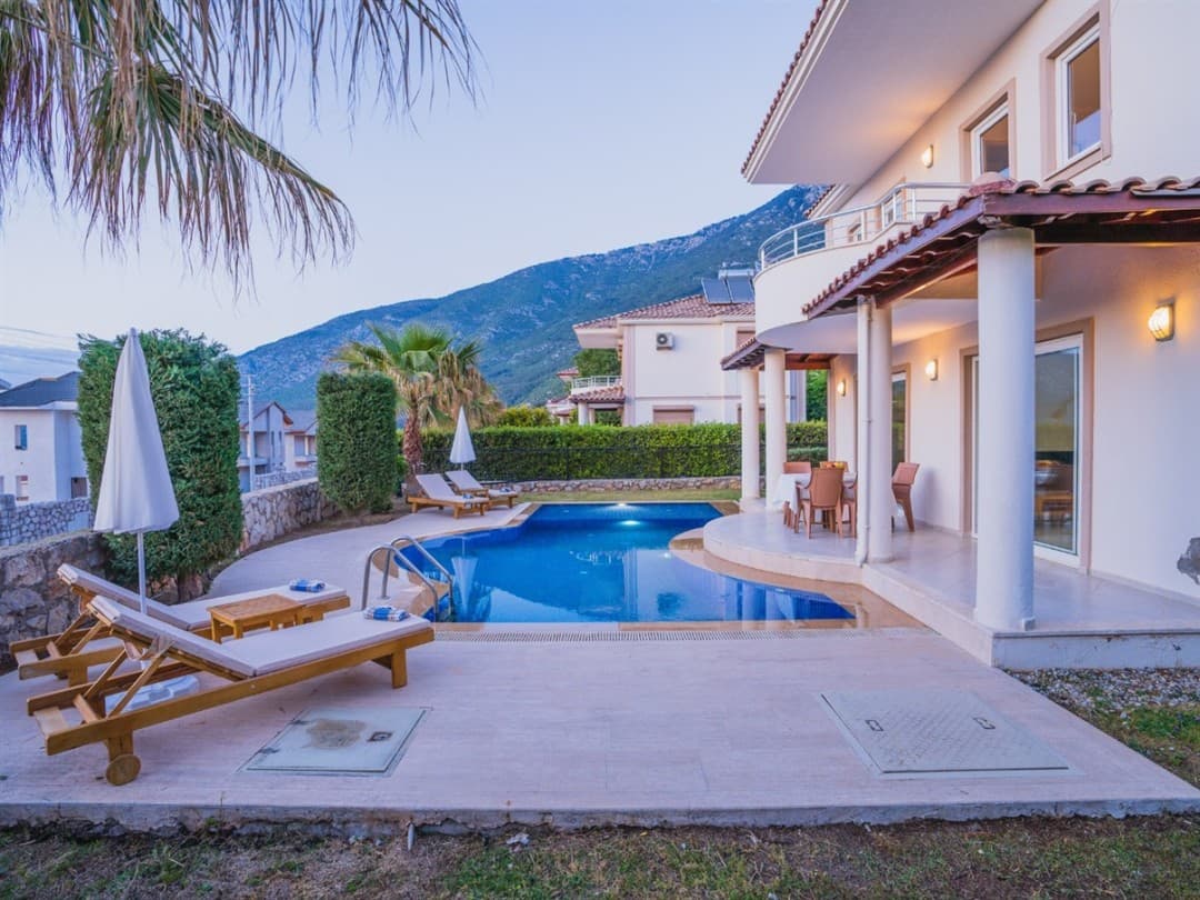 Gündoğumunda havuz kenarında şezlonglar ve arka planda dağ manzaralı villa.