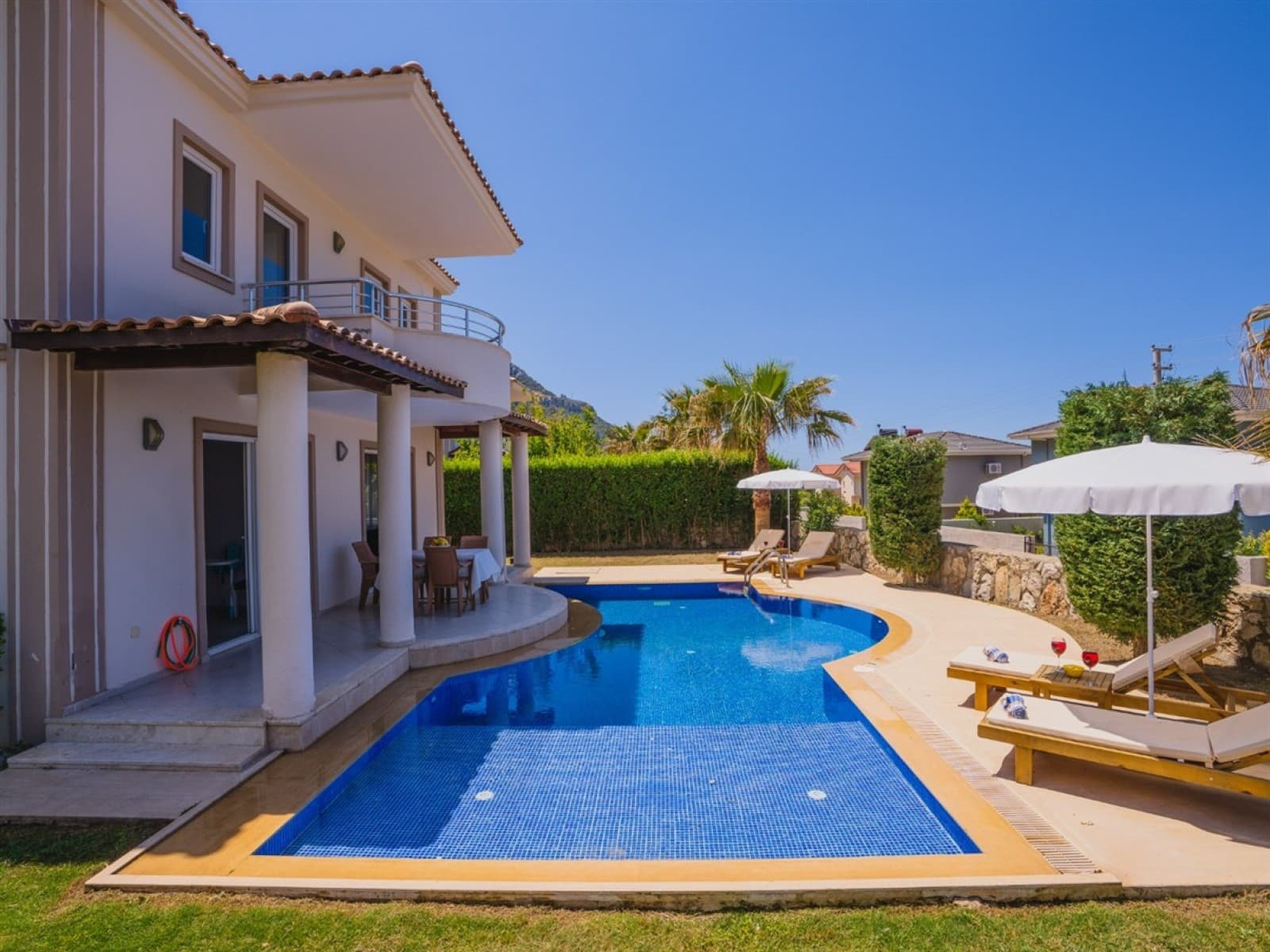İki katlı beyaz villa, kavisli yüzme havuzu, şezlonglar ve şemsiyeler.