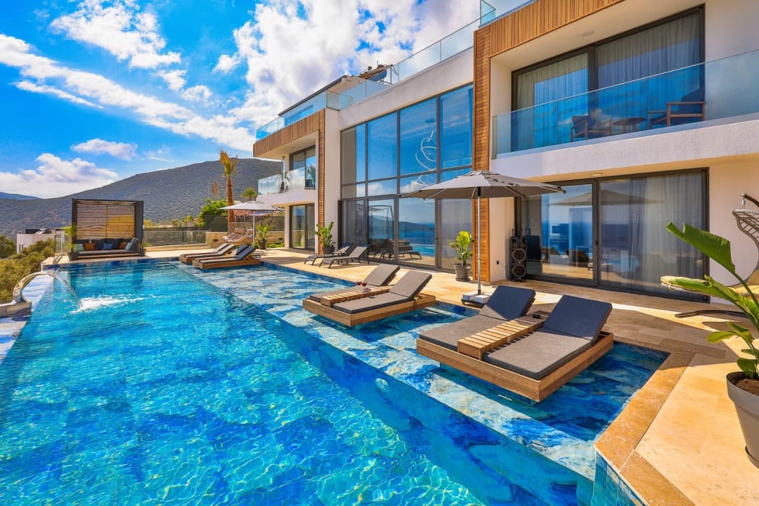Gündüz havuz kenarında şezlonglar ve arka planda dağ manzarası olan modern villa.