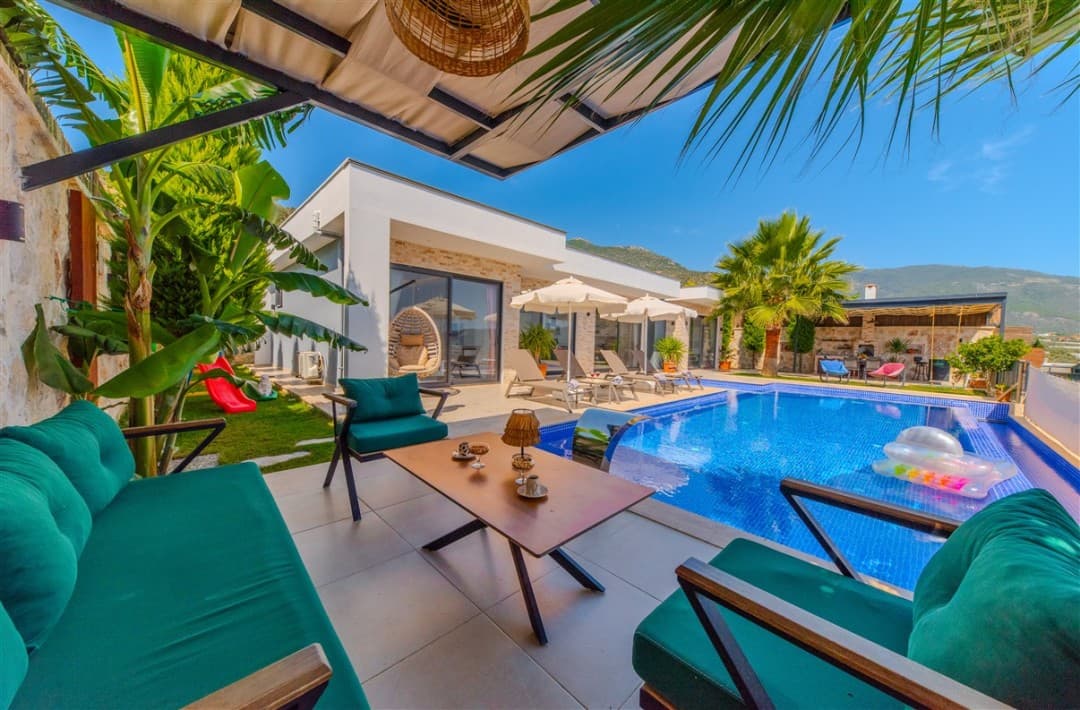 Ön planda yeşil minderli koltuklar ve sehpa, arkasında havuz ve beyaz dış cepheli villa yer alıyor.
