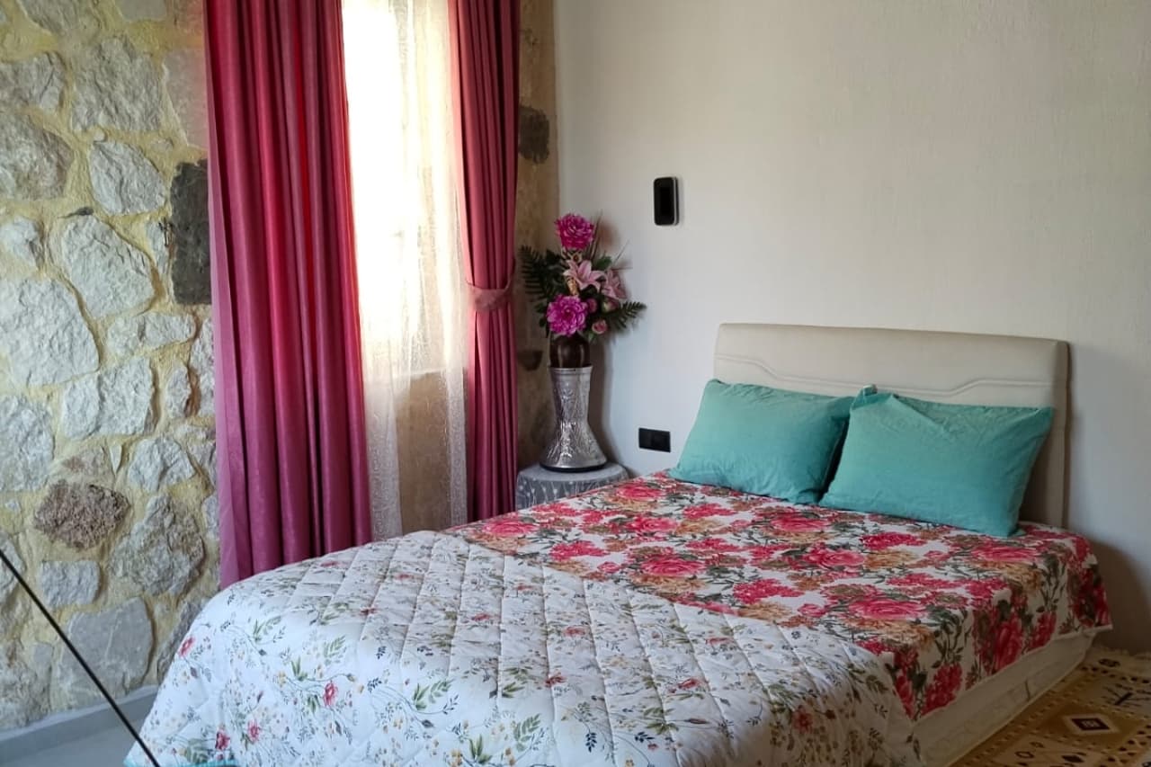 Taş duvarlı yatak odasında pembe perdeler, çiçekler ve desenli yatak örtüsü yer alıyor.