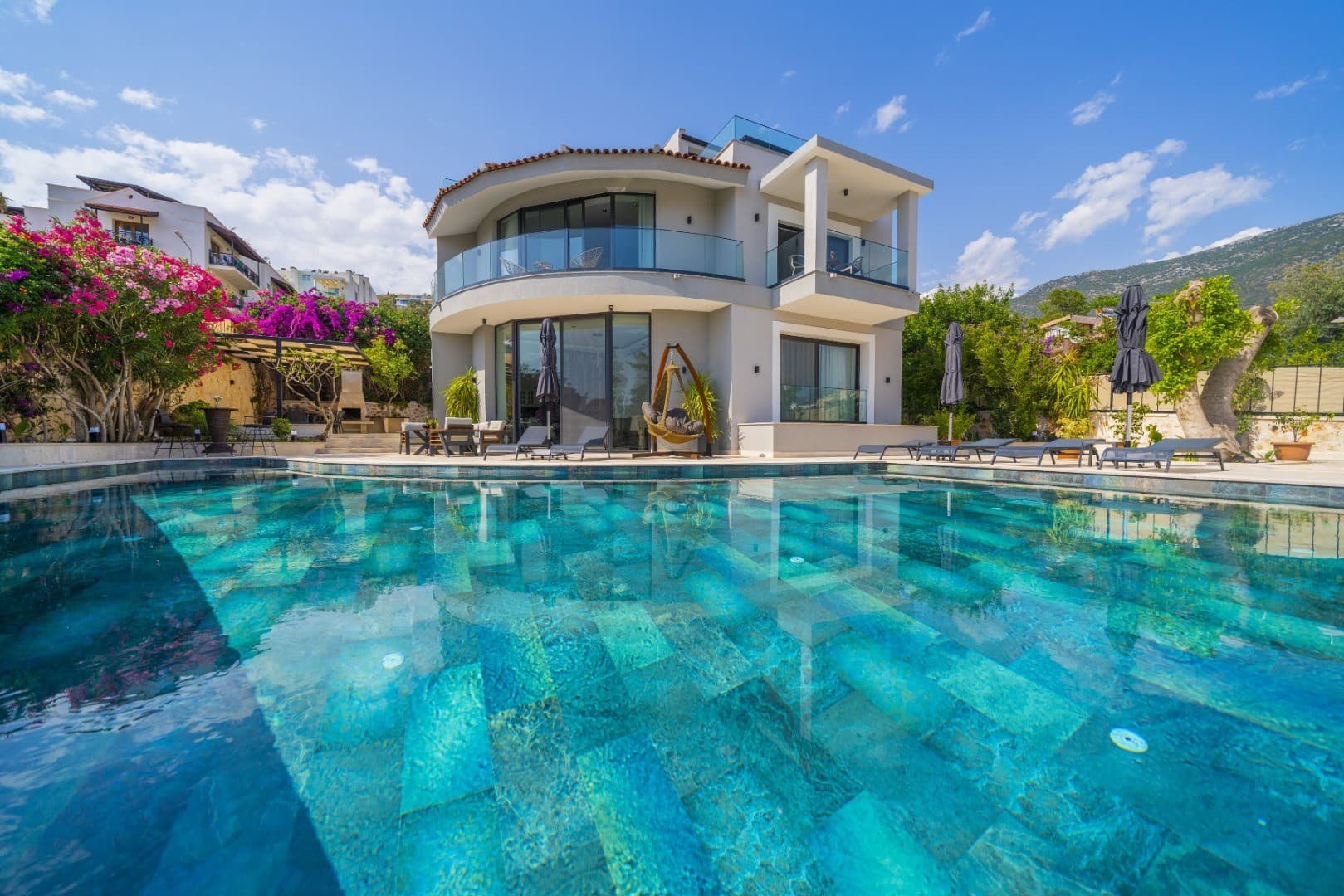 Ön planda turkuaz renkli bir yüzme havuzu, arka planda ise balkonlu, modern iki katlı bir villa bulunuyor.