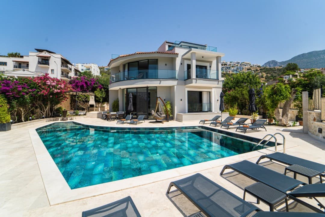 Açık havuzlu iki katlı modern villa, önünde şezlonglar ve arka planda dağ manzarası.