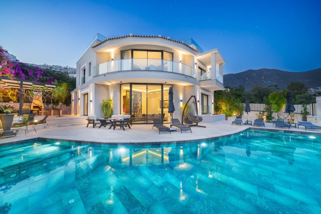 Gece ışıklandırmalı kavisli havuzu ve şezlongları olan lüks villa.