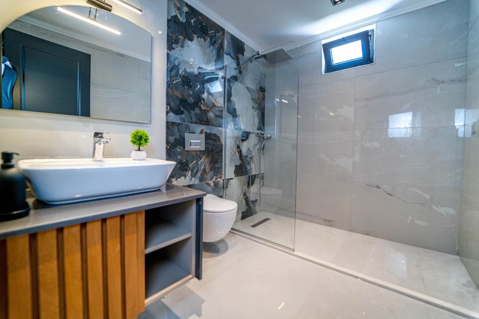 Modern banyoda ahşap dolaplı lavabo, siyah beyaz mermer duvarlı cam duşakabin ve asma klozet.