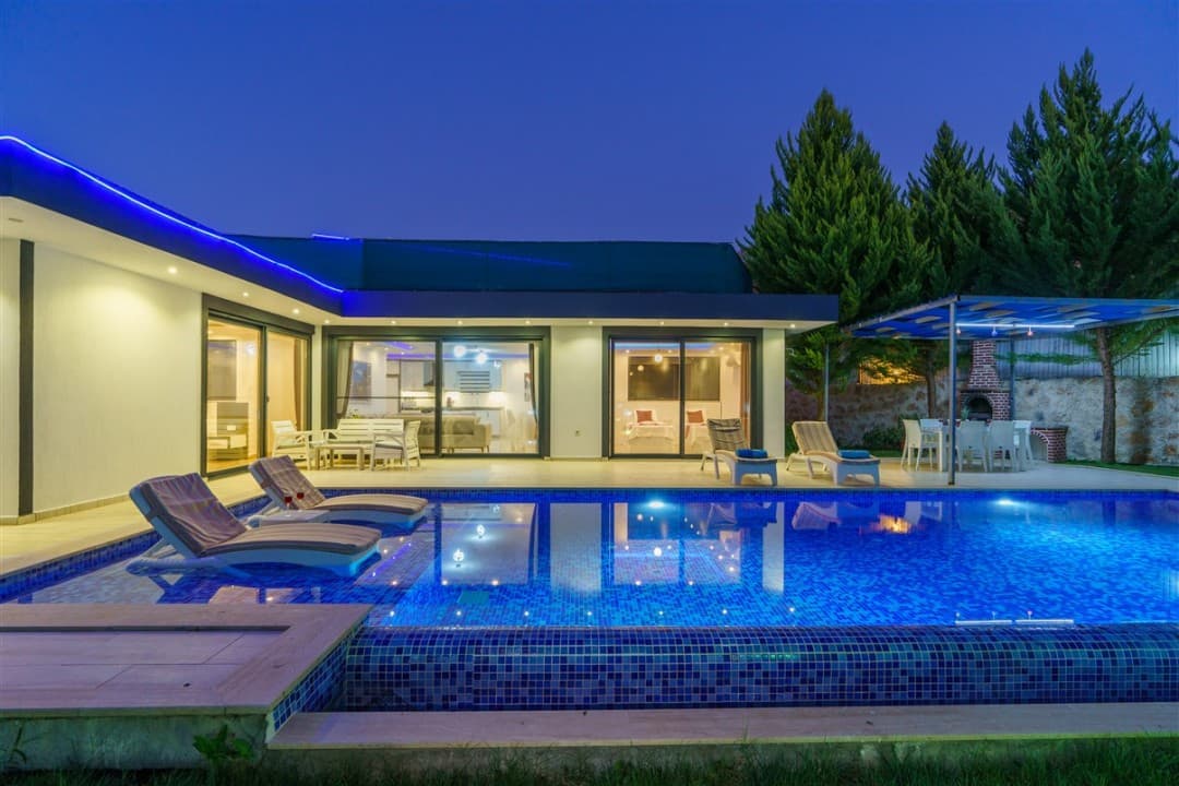 Gece aydınlatmalı özel havuzu, şezlongları ve veranda oturma grubu bulunan modern tek katlı villa.