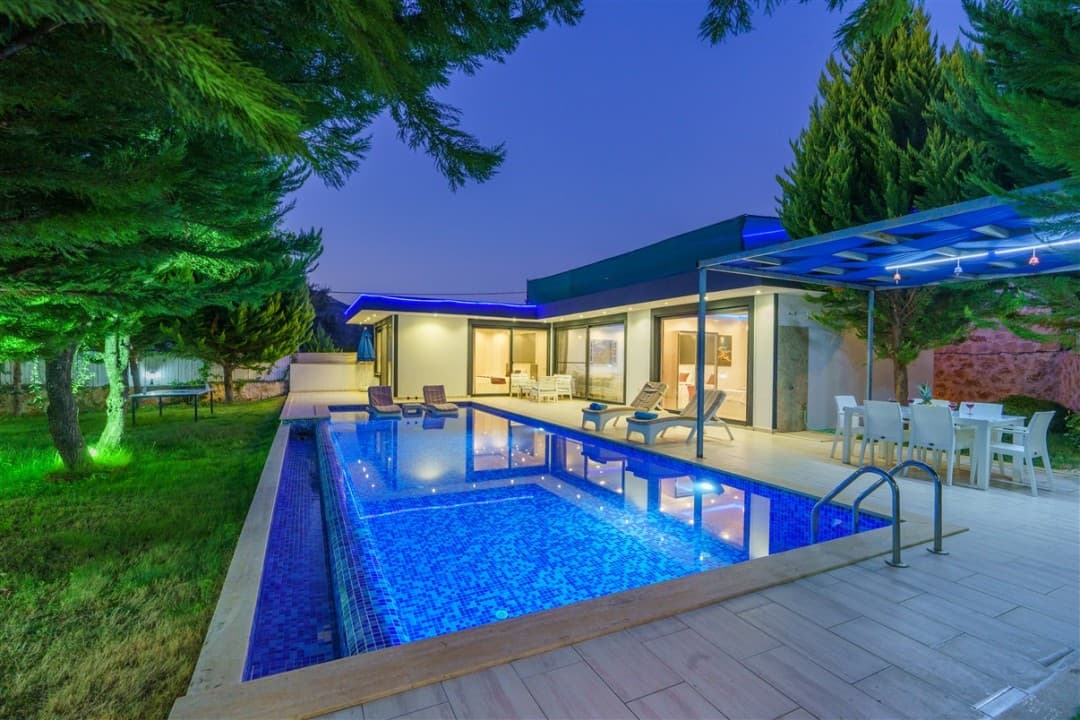 Akşam vakti havuz başı, şezlonglar ve yemek masası olan tek katlı villa.