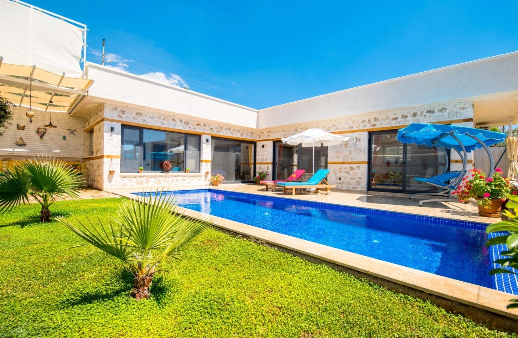 Çim bahçesi, havuzu ve mavi şezlongları olan tek katlı taş villa.