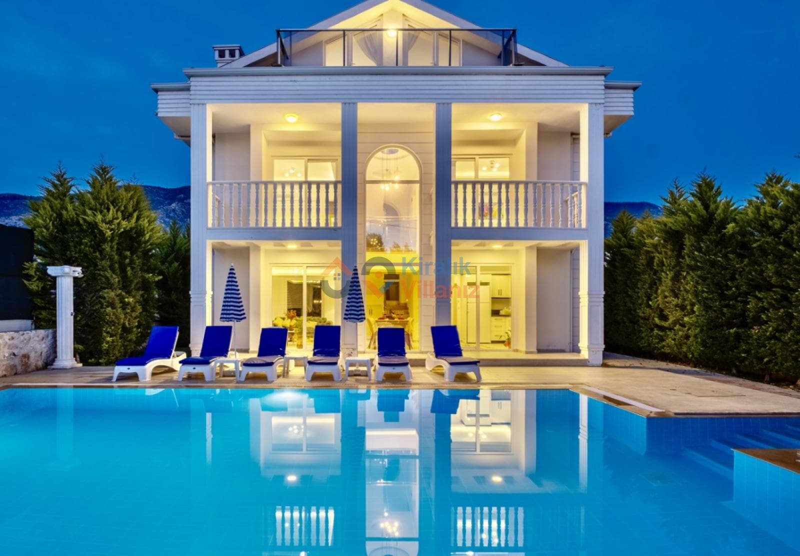 Akşam aydınlatmalı havuzu ve şezlongları bulunan iki katlı villa.