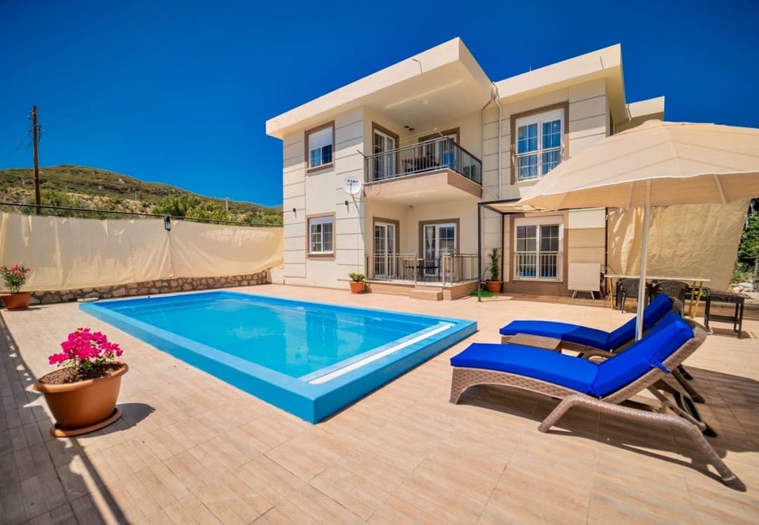 Mavi havuzlu, iki katlı bej renkli villa, güneş şemsiyesi, şezlonglar ve saksı çiçekleri.