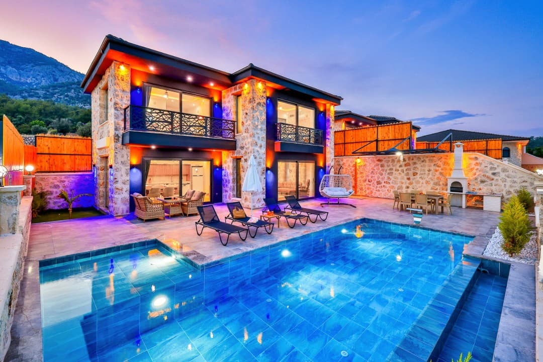 Gün batımında havuzlu, taş kaplamalı iki katlı villa ve bahçesinde şezlonglar, oturma grubu ile yemek masası.