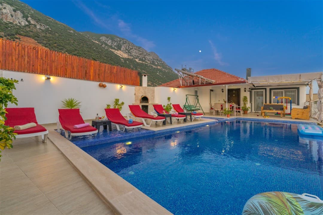 Dağ manzaralı, havuz kenarında kırmızı şezlonglar ve taş fırın bulunan havuzlu villa bahçesi.