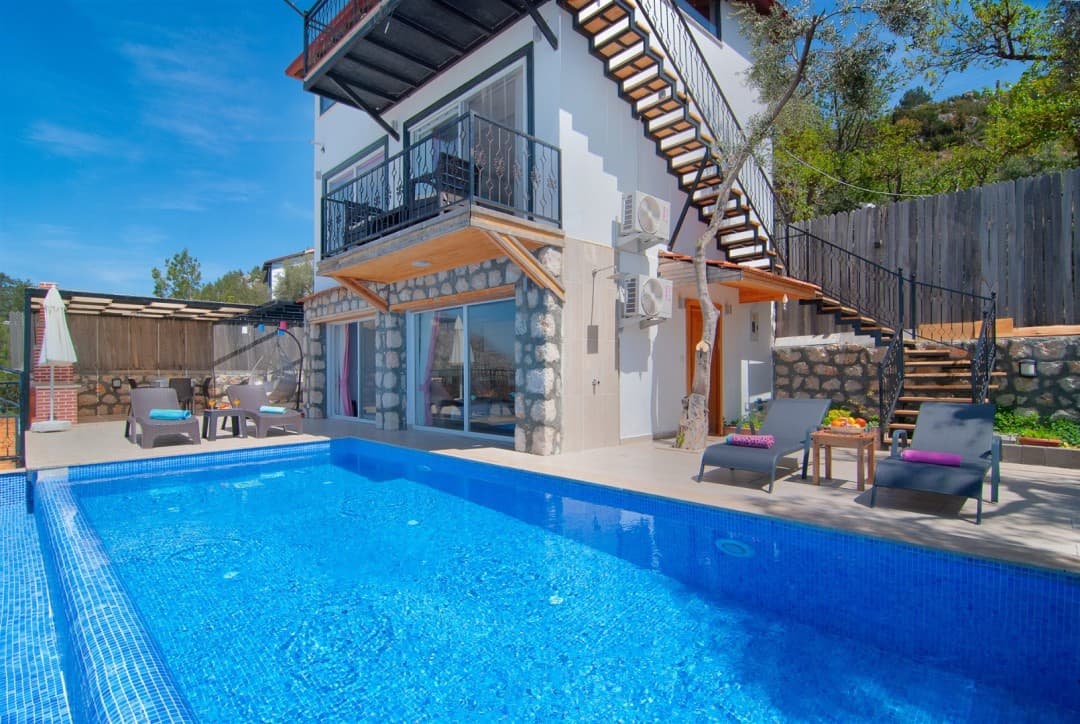 Havuz kenarında şezlonglar ve dış merdiveni olan beyaz duvarlı taş detaylı villa.