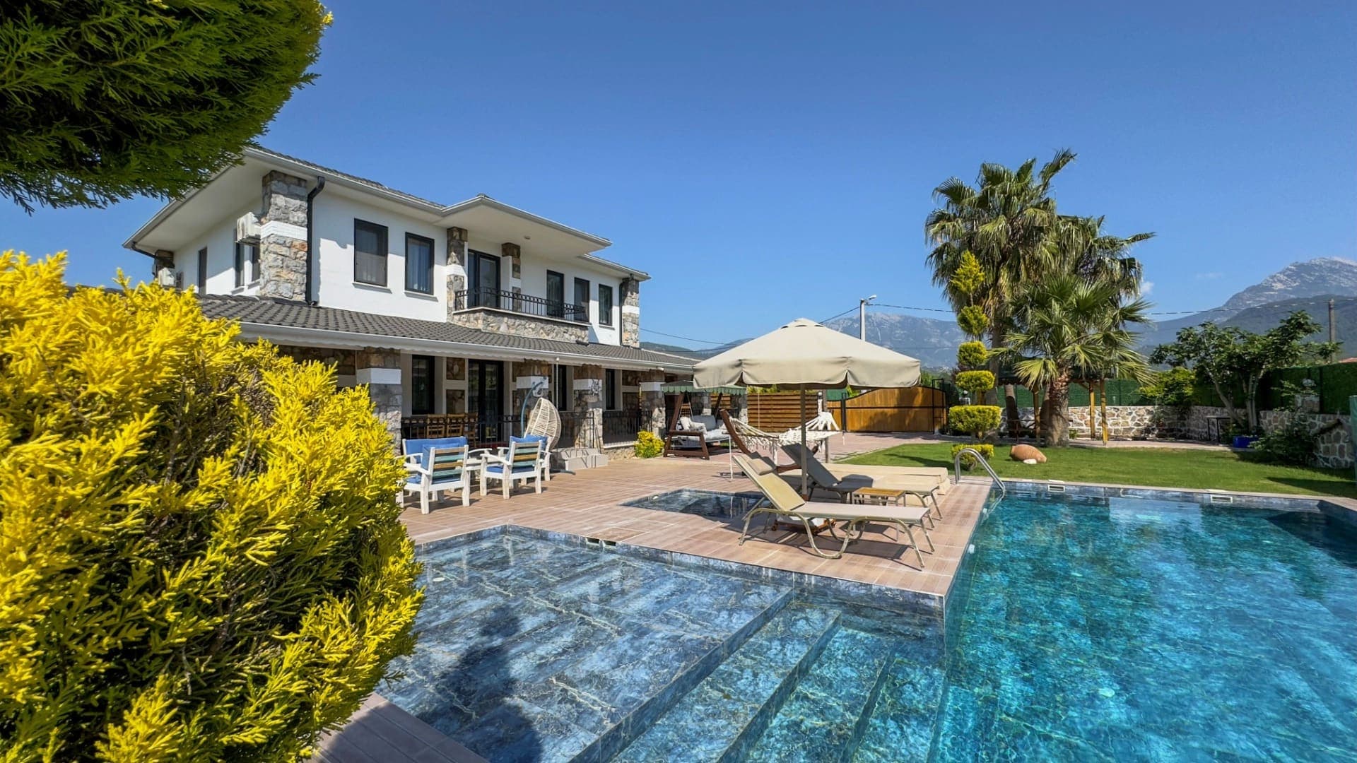 Bahçesinde havuz, şezlonglar ve şemsiyeler bulunan iki katlı taş detaylı villa.