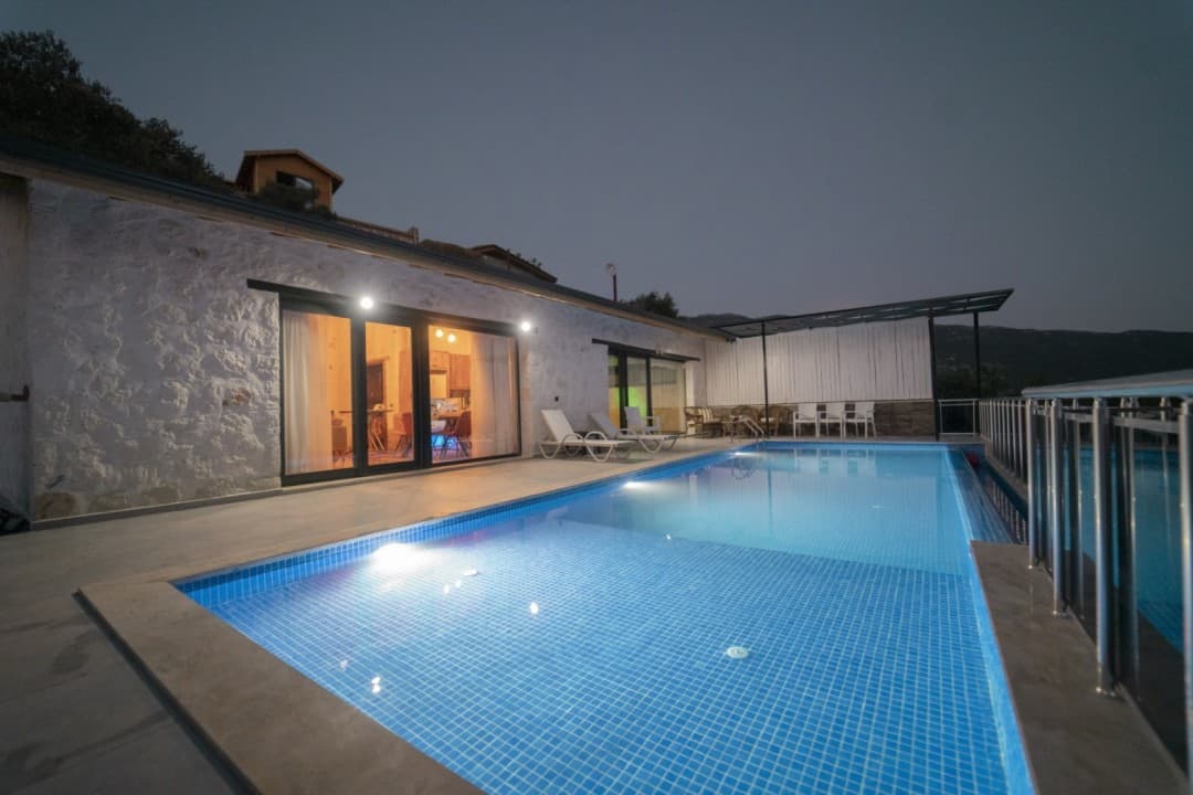 Gece aydınlatmalı yüzme havuzu ve önünde şezlonglar bulunan taş duvarlı villa dış cephesi.