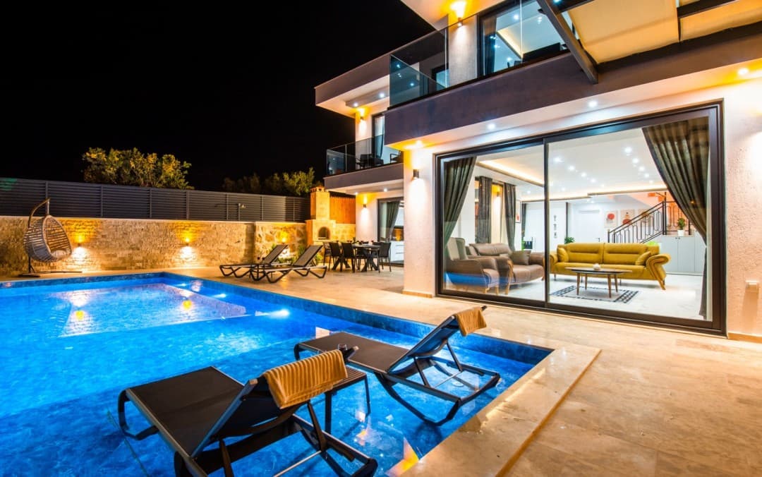 Geceleri aydınlatmalı havuzu ve şezlongları bulunan iki katlı modern villanın dış görünümü.