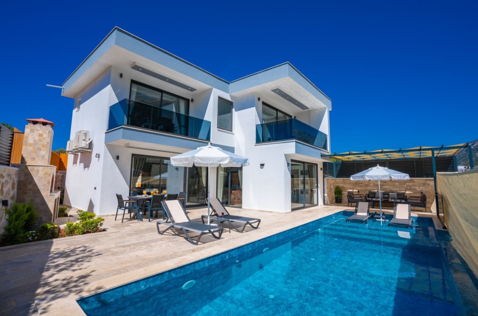 Mavi gökyüzü altında beyaz villa, önünde havuz ve şezlonglar bulunuyor.