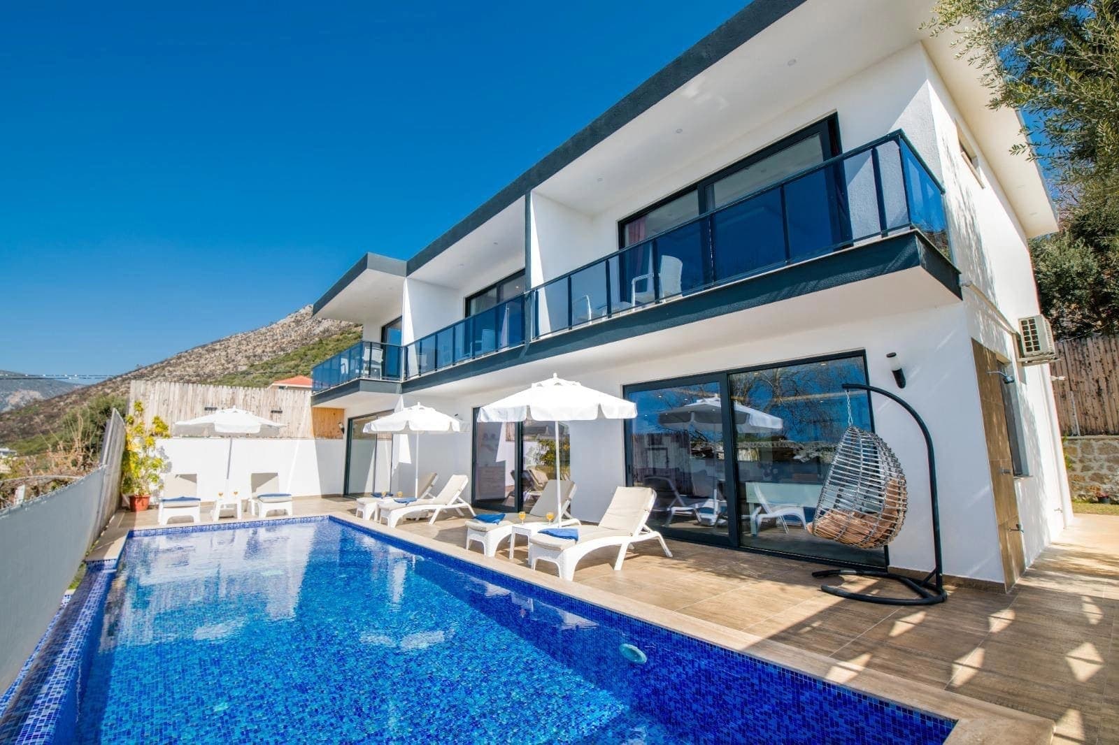 Önünde havuz, şezlonglar ve asma salıncak bulunan beyaz dış cepheli villa.