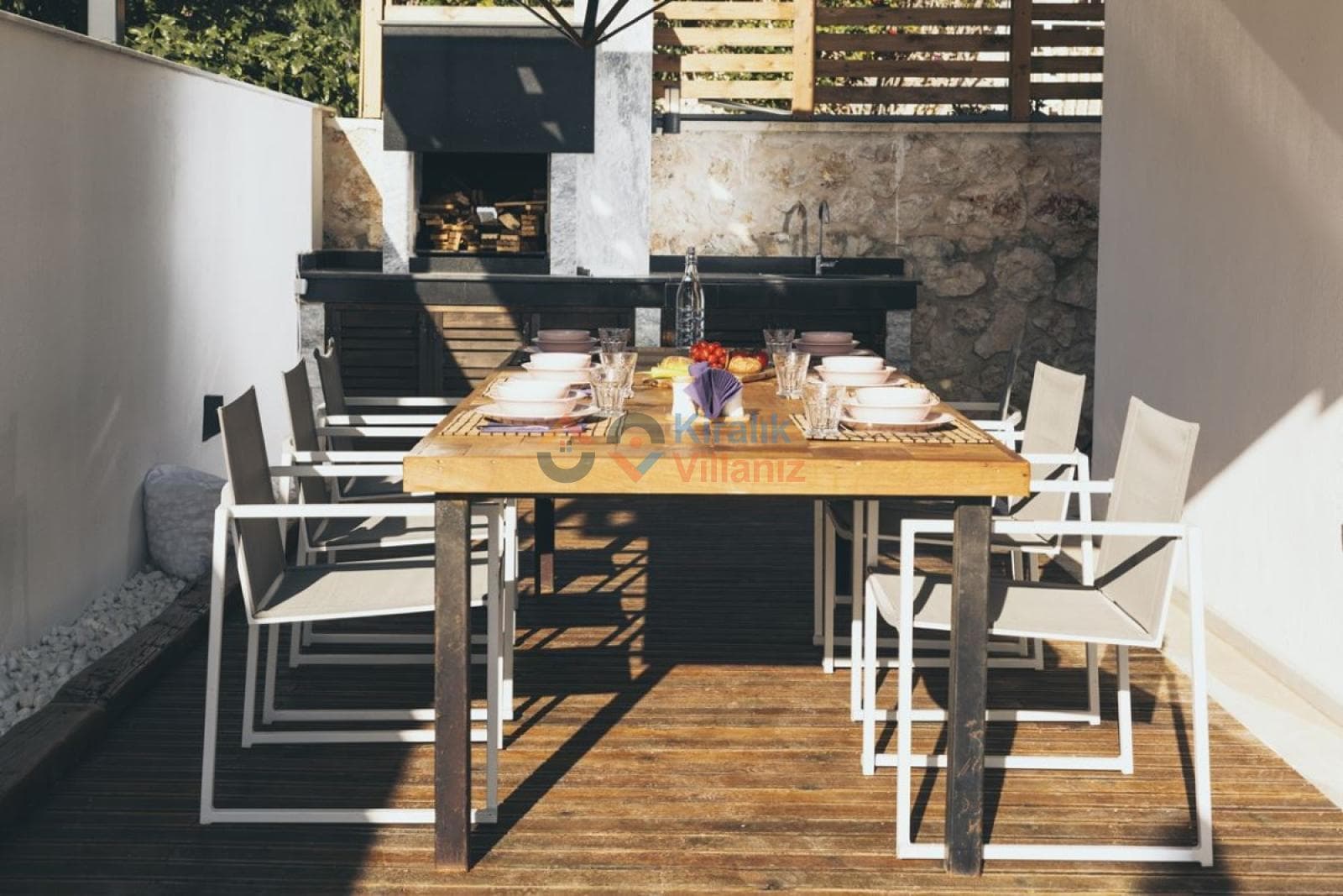 Ahşap yemek masası, beyaz sandalyeler ve arka planda barbekü alanı.