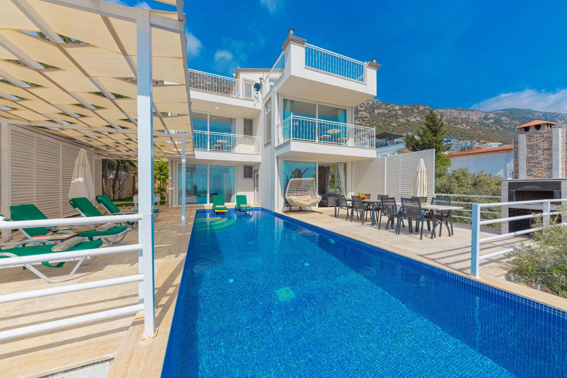 Mavi havuzlu, şezlonglu ve dağ manzaralı villa dış cephesi.