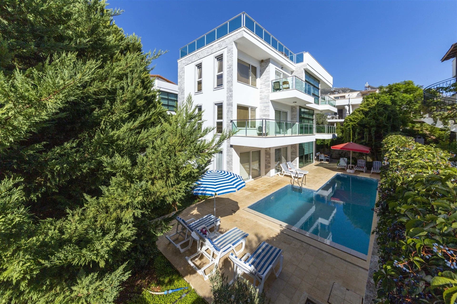 Ağaçlarla çevrili havuzlu ve şezlonglu lüks villa dış cephesi.