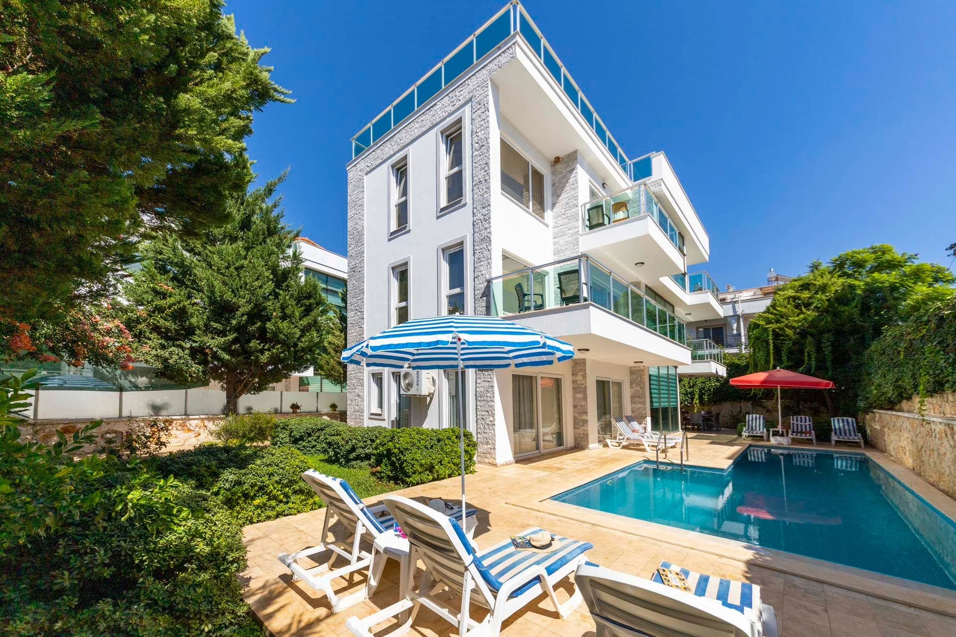 Güneş şemsiyeli şezlonglar ve havuzun bulunduğu villa bahçesi.