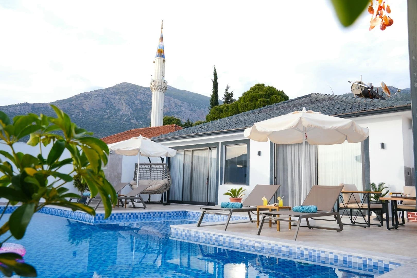 Arka planda dağ ve cami minaresi olan, havuz kenarında şezlong ve şemsiyeler bulunan villa dış cephesi.