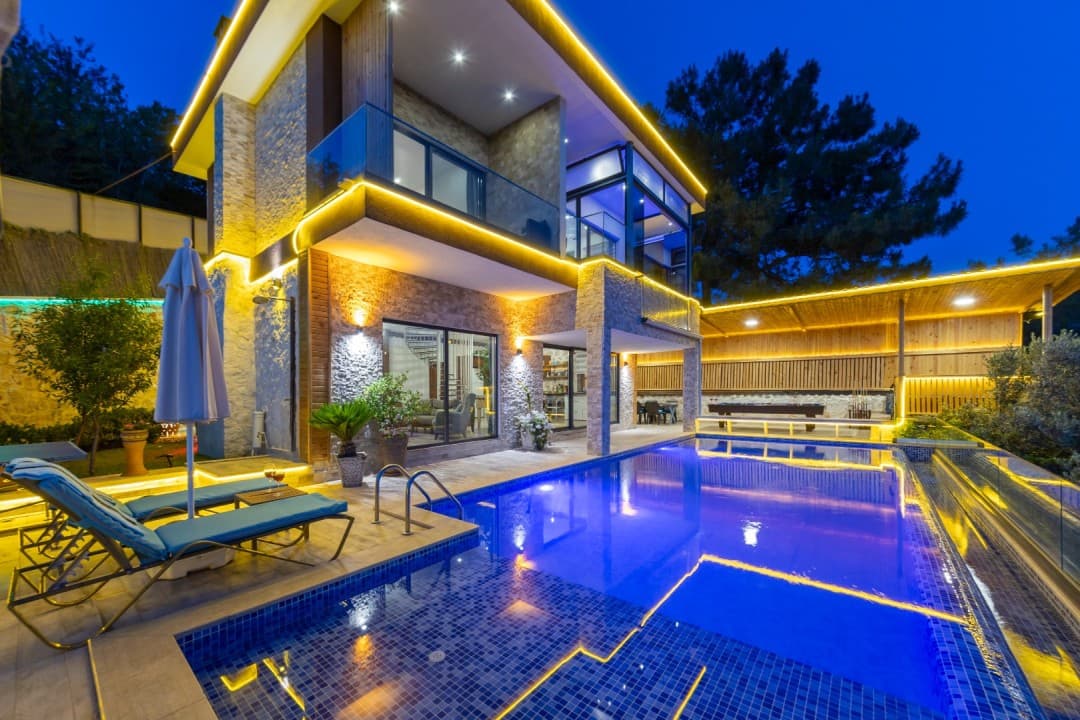 Gece aydınlatmalı, havuz başında şezlong ve şemsiye bulunan taş kaplamalı iki katlı villa.