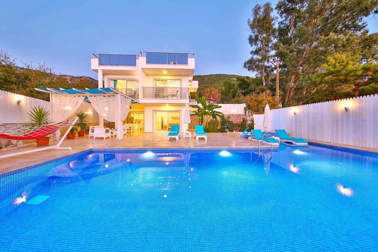 Aydınlatmalı havuz kenarında şezlonglar ve beyaz cepheli villa