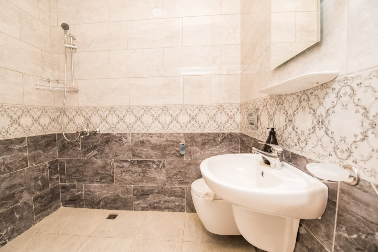 Gri ve bej desenli seramik duvarlara sahip, lavabo ve duş başlığı görünen banyo.
