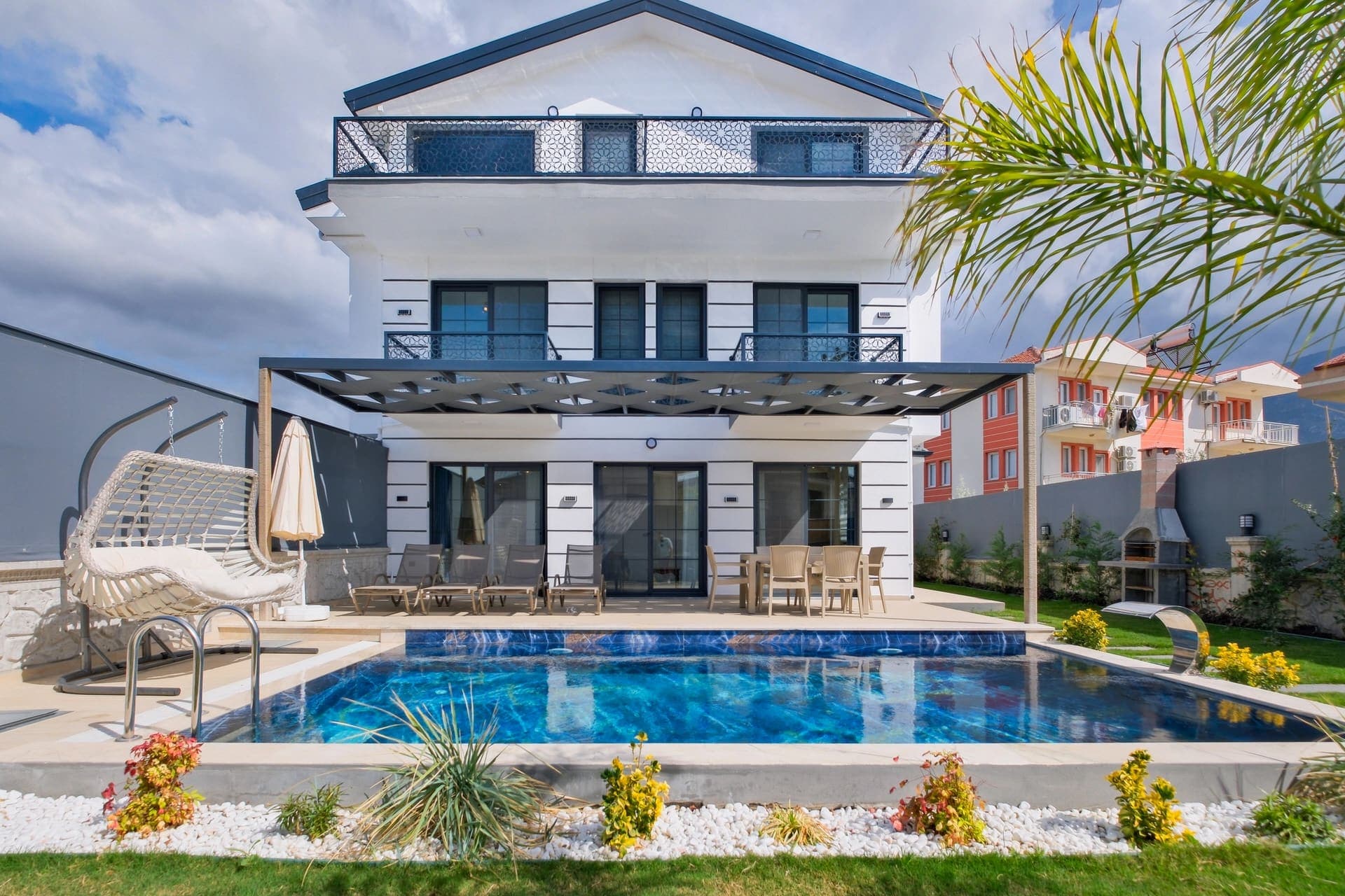 Önünde yüzme havuzu, şezlonglar ve salıncak bulunan beyaz villa dış cephesi.