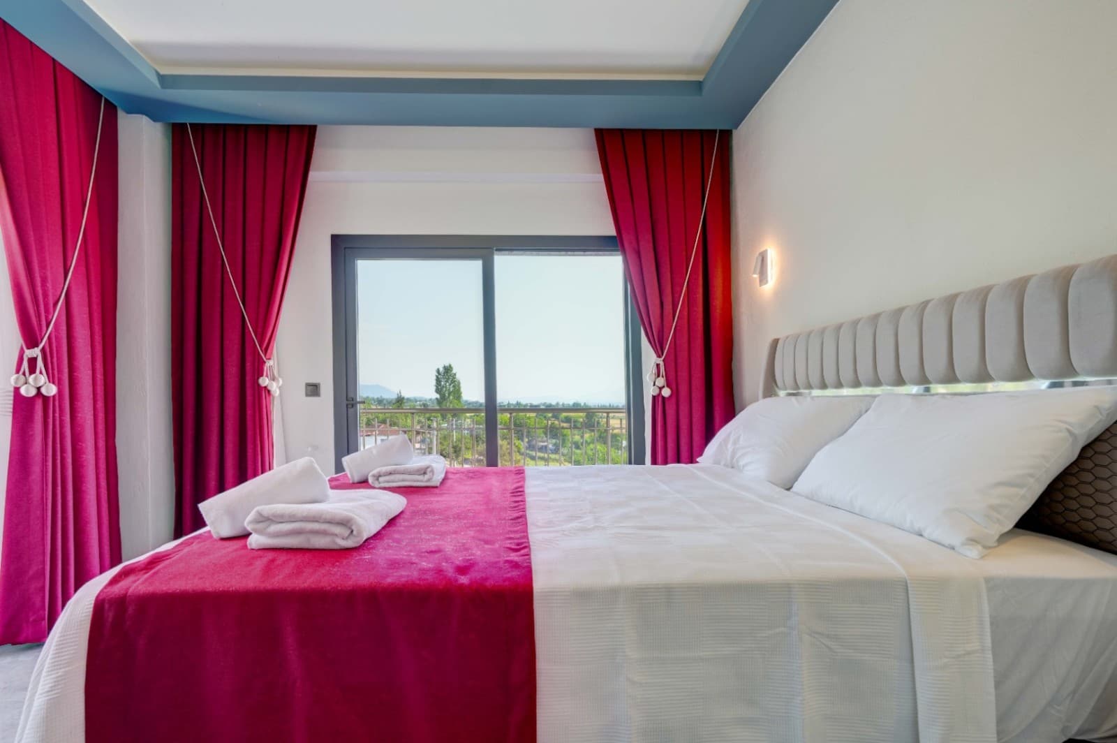 Kırmızı perdeli ve manzaralı cam balkon kapısının önünde beyaz çift kişilik yatak.